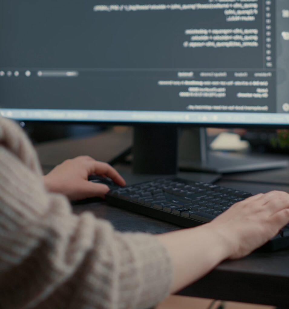 Hände auf einer Tastatur vor einem Bildschirm mit Programmcode